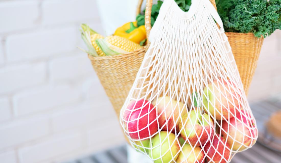 Frau hält Strohkorb und wiederverwendbare Mesh-Einkaufstasche mit Äpfeln, Gemüse, weißen Backstein Hintergrund. 