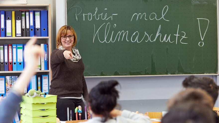 Klassenzimmer mit Schülern und Lehrerin. Auf der Tafel steht "Probier mal Klimaschutz"