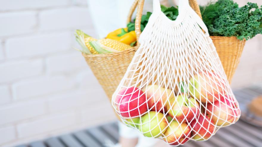 Frau hält Strohkorb und wiederverwendbare Mesh-Einkaufstasche mit Äpfeln, Gemüse, weißen Backstein Hintergrund. 