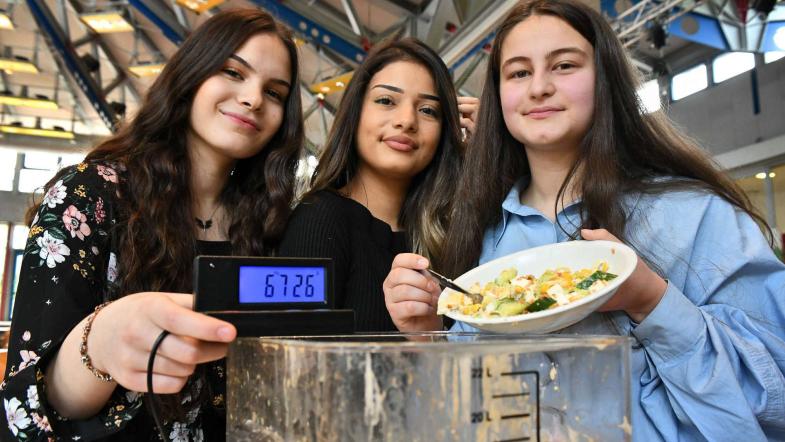 Drei Schülerinnen blicken in die Kamera, vor ihnen eine Waage, mit der sie ihre Lebensmittelabfälle wiegen