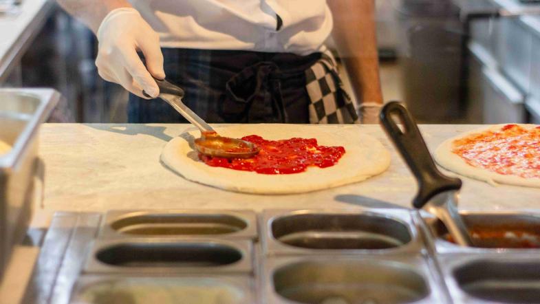 Ein Mann verteilt Tomatensauce auf einem ausgerollten Pizzateig.